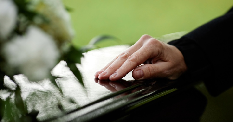 Mão toca carinhosamente a superfície de uma urna envernizada em processos funerários.